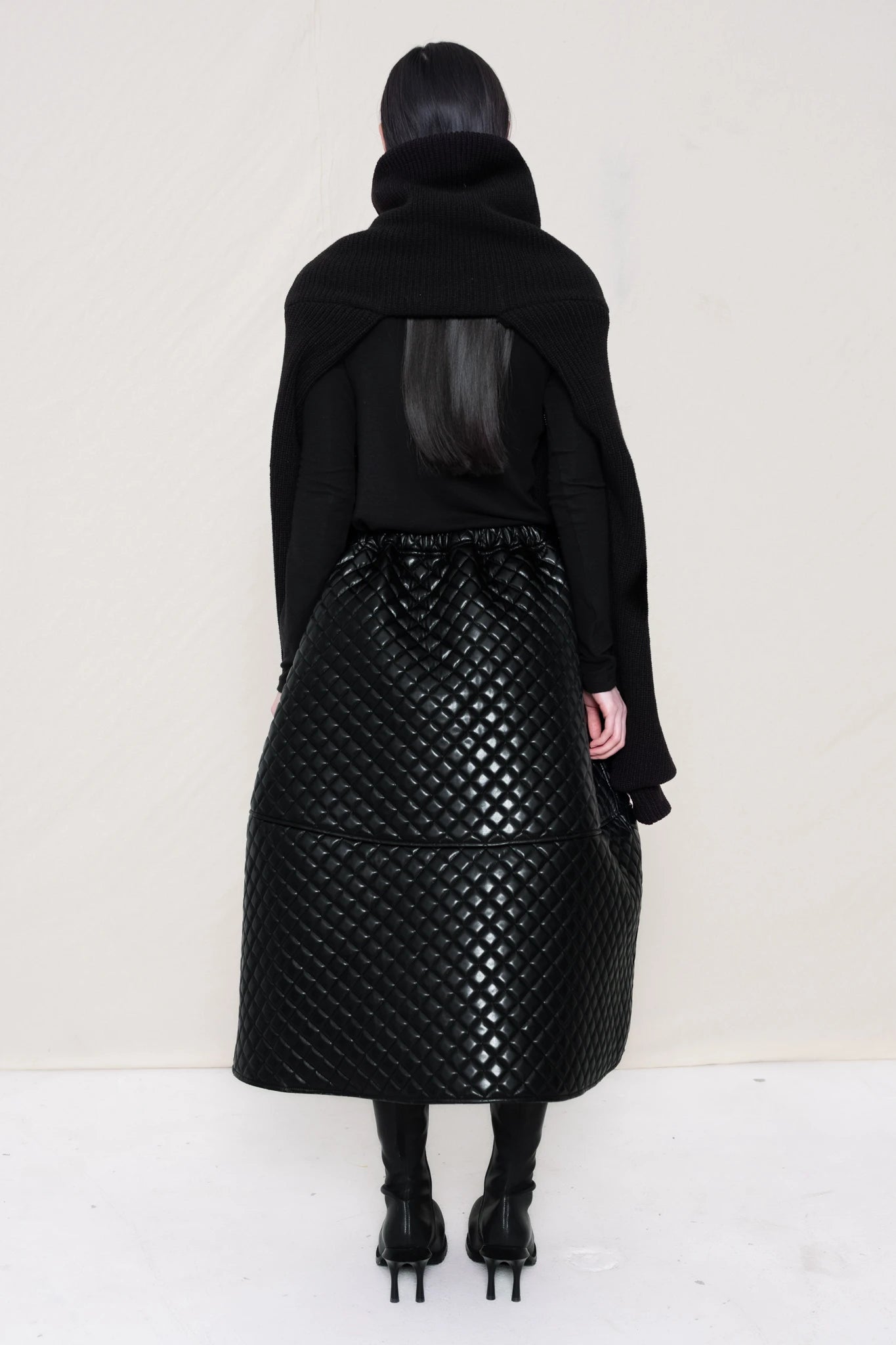 PERSON-WEARING-BLACK-CHUNKY-KNIT-TURTLENECK-THROW-OVER-WITH-BLACK-QUILTED-A-LINE-SKIRT-AND-BLACK-COWHIDE-DOUBLE-HEELED-BOOTS-HOLDING-BLACK-A-LINE-SHOPPER-WITH-PIERCING-DETAIL-BACK-VIEW