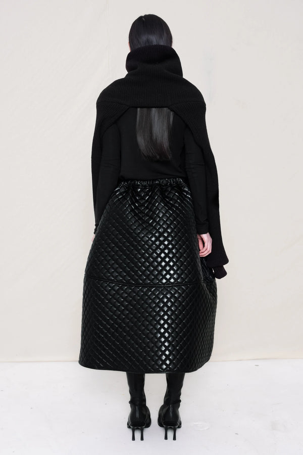 PERSON-WEARING-BLACK-CHUNKY-KNIT-TURTLENECK-THROW-OVER-WITH-BLACK-QUILTED-A-LINE-SKIRT-AND-BLACK-COWHIDE-DOUBLE-HEELED-BOOTS-HOLDING-BLACK-A-LINE-SHOPPER-WITH-PIERCING-DETAIL-BACK-VIEW