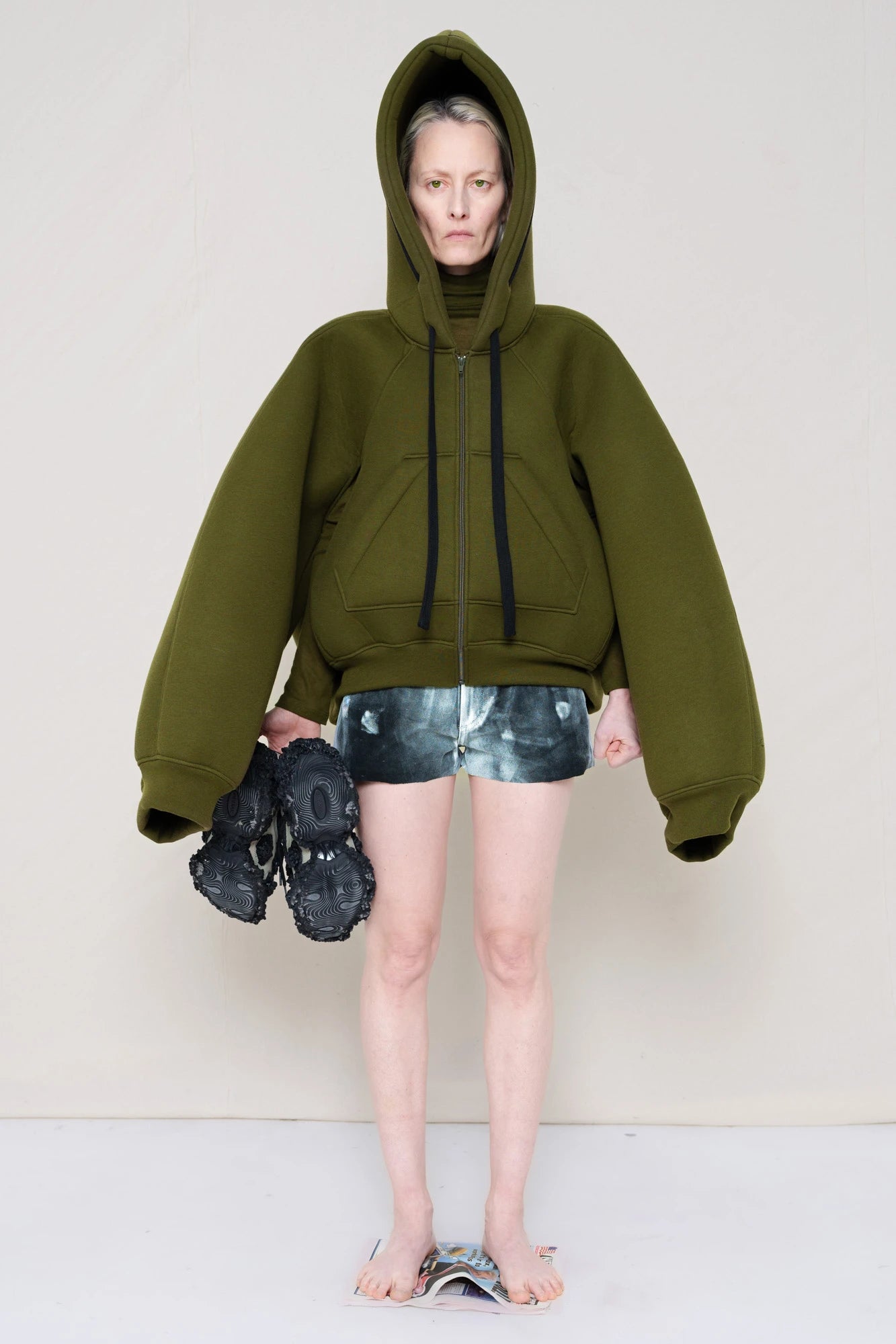 PERSON-WEARING-EXTREMELY-FOAMY-HOODIE-IN-THE-COLOR-MOSS-PAIRED-WITH-PAINTED-DENIM-SHORTS-WHILE-HOLDING-CUSTOMIZED-SNEAKERS-AND-STANDING-ON-NEWSPAPER-FRONT-VIEW