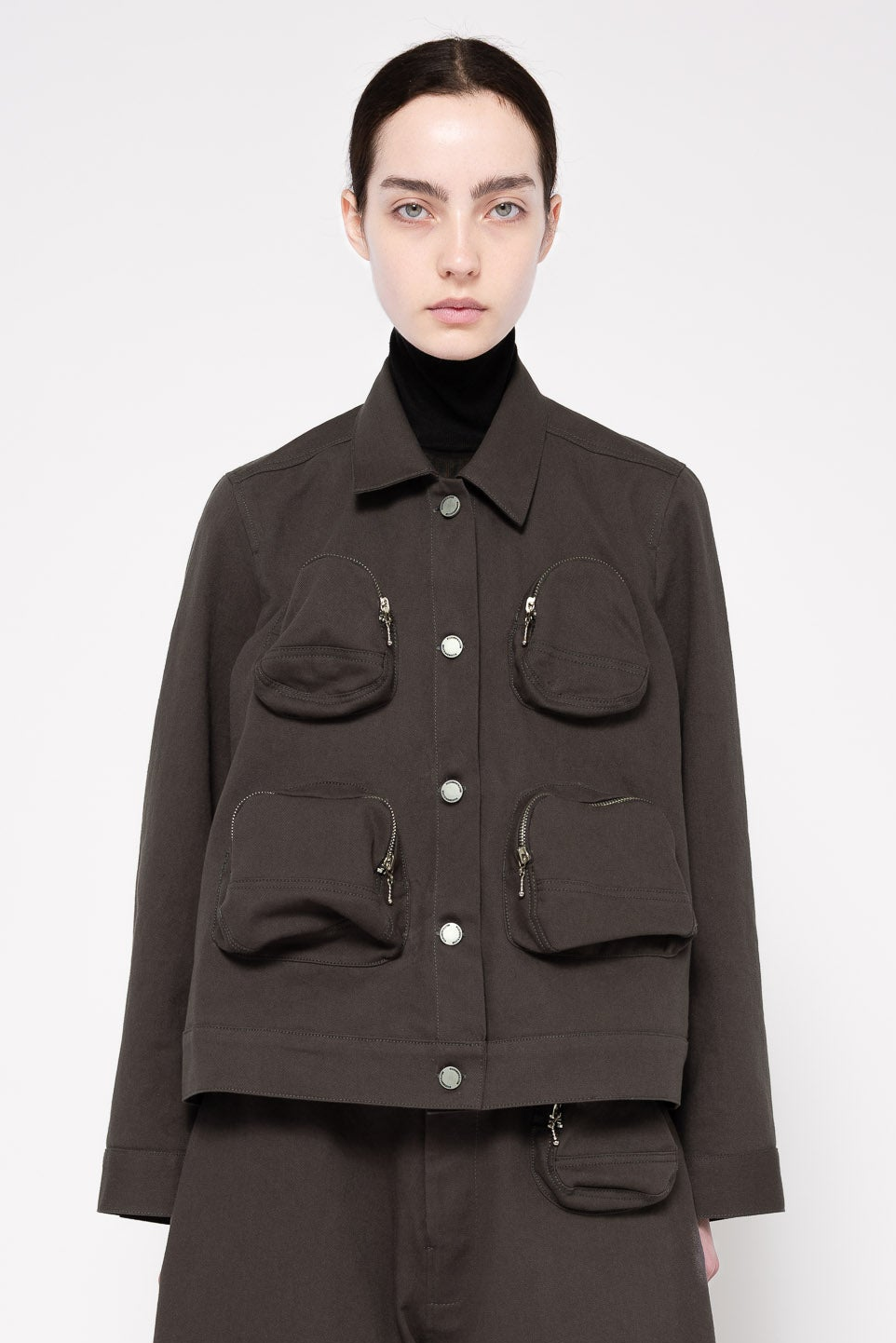 A model wearing a dark grey utility jacket with multiple front pockets and silver-toned hardware, styled over a black turtleneck sweater against a light gray studio background.