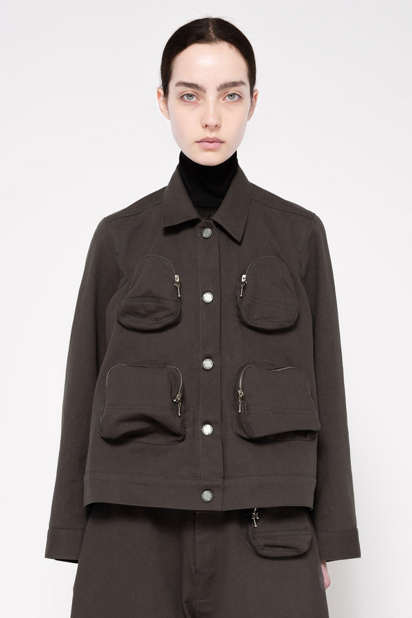 A model wearing a dark grey utility jacket with multiple front pockets and silver-toned hardware, styled over a black turtleneck sweater against a light gray studio background.