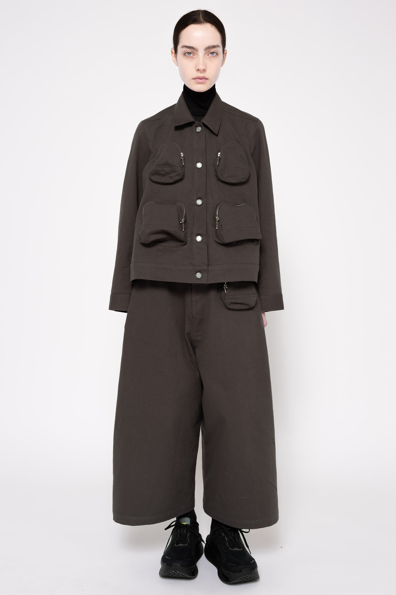 A model stands against a light gray studio background wearing a dark grey cropped utility jacket with front flap pockets, matching wide-leg trousers, a black turtleneck, and black shoes.