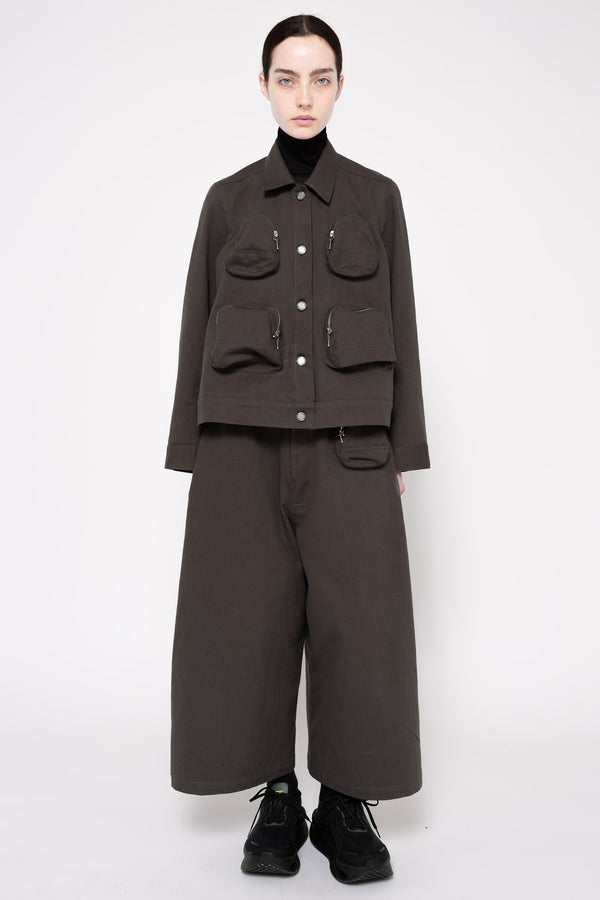 A model stands against a light gray studio background wearing a dark grey cropped utility jacket with front flap pockets, matching wide-leg trousers, a black turtleneck, and black shoes.