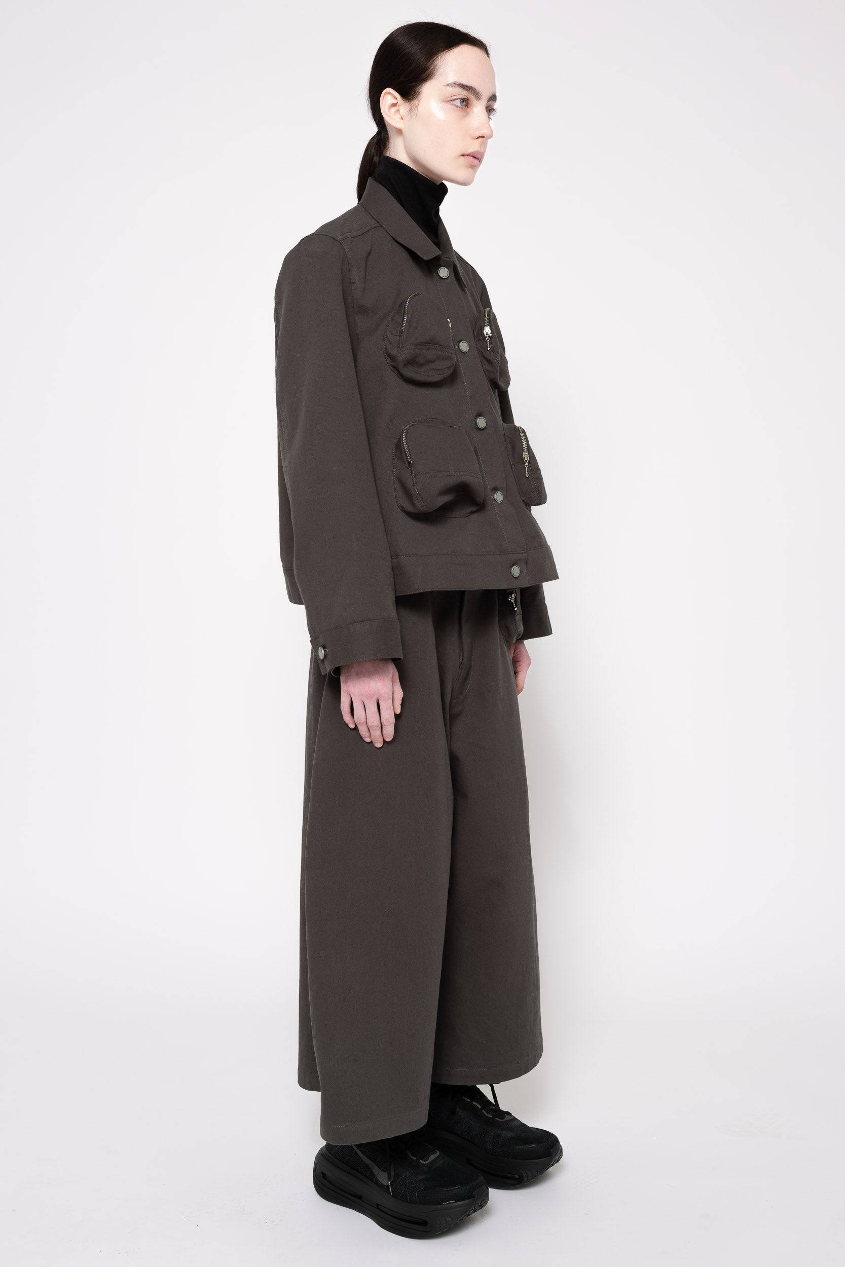 A model is shown in profile wearing a dark grey cropped utility jacket with multiple flap pockets, matching wide-leg trousers, a black turtleneck, and black shoes, set against a plain light gray studio background.