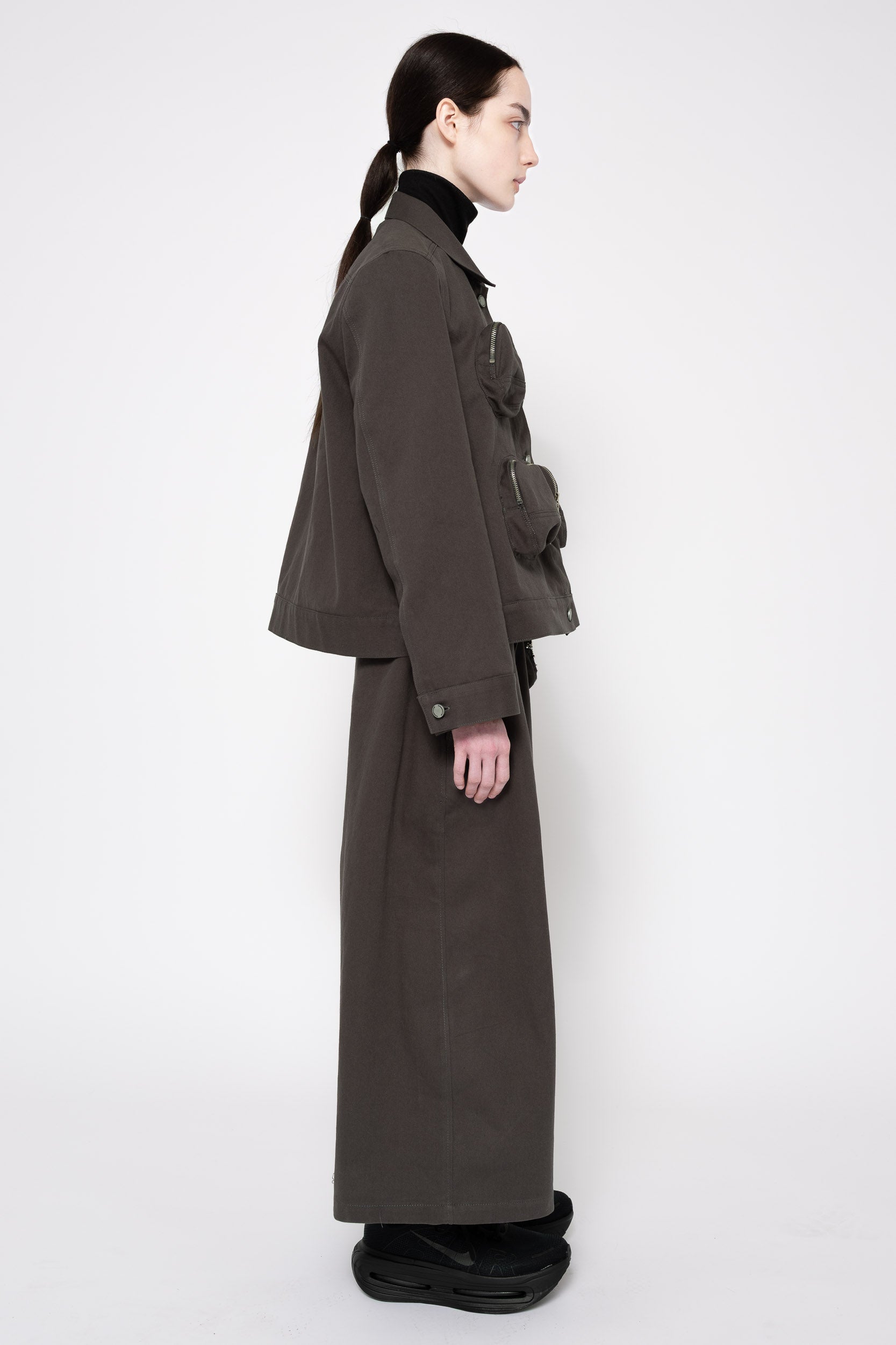 A model shown in profile wearing a dark grey utility jacket and matching wide-leg skirt, styled with a black turtleneck and black platform shoes against a plain white studio background.