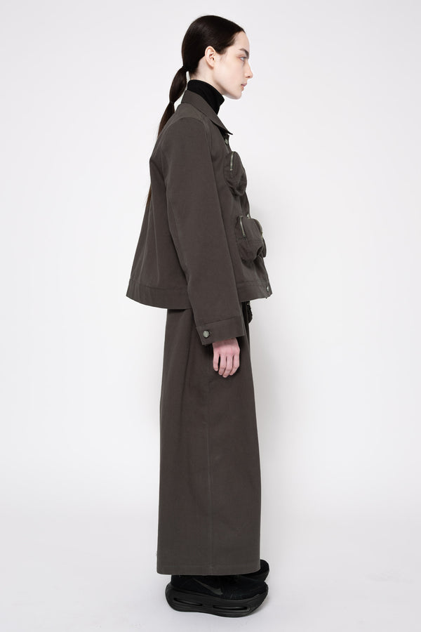 A model shown in profile wearing a dark grey utility jacket and matching wide-leg skirt, styled with a black turtleneck and black platform shoes against a plain white studio background.