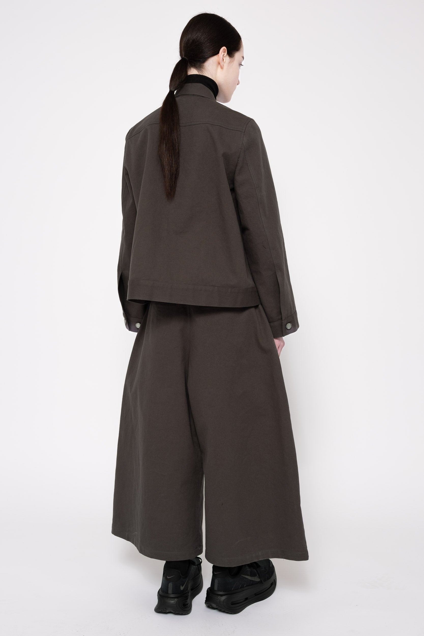 A model shown from the back wearing a dark grey cropped jacket and matching wide-leg trousers, paired with black boots, against a plain white studio background.