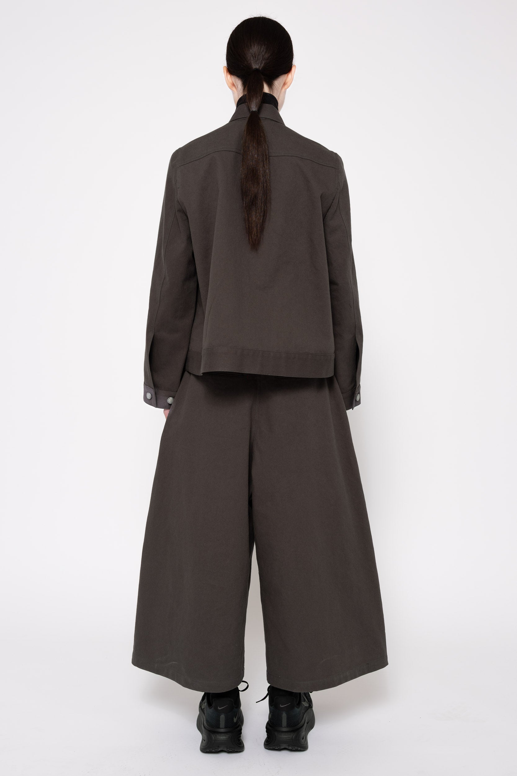 A model shown from the back wearing a dark grey cropped jacket and matching wide-leg trousers, paired with black boots, against a plain white studio background.