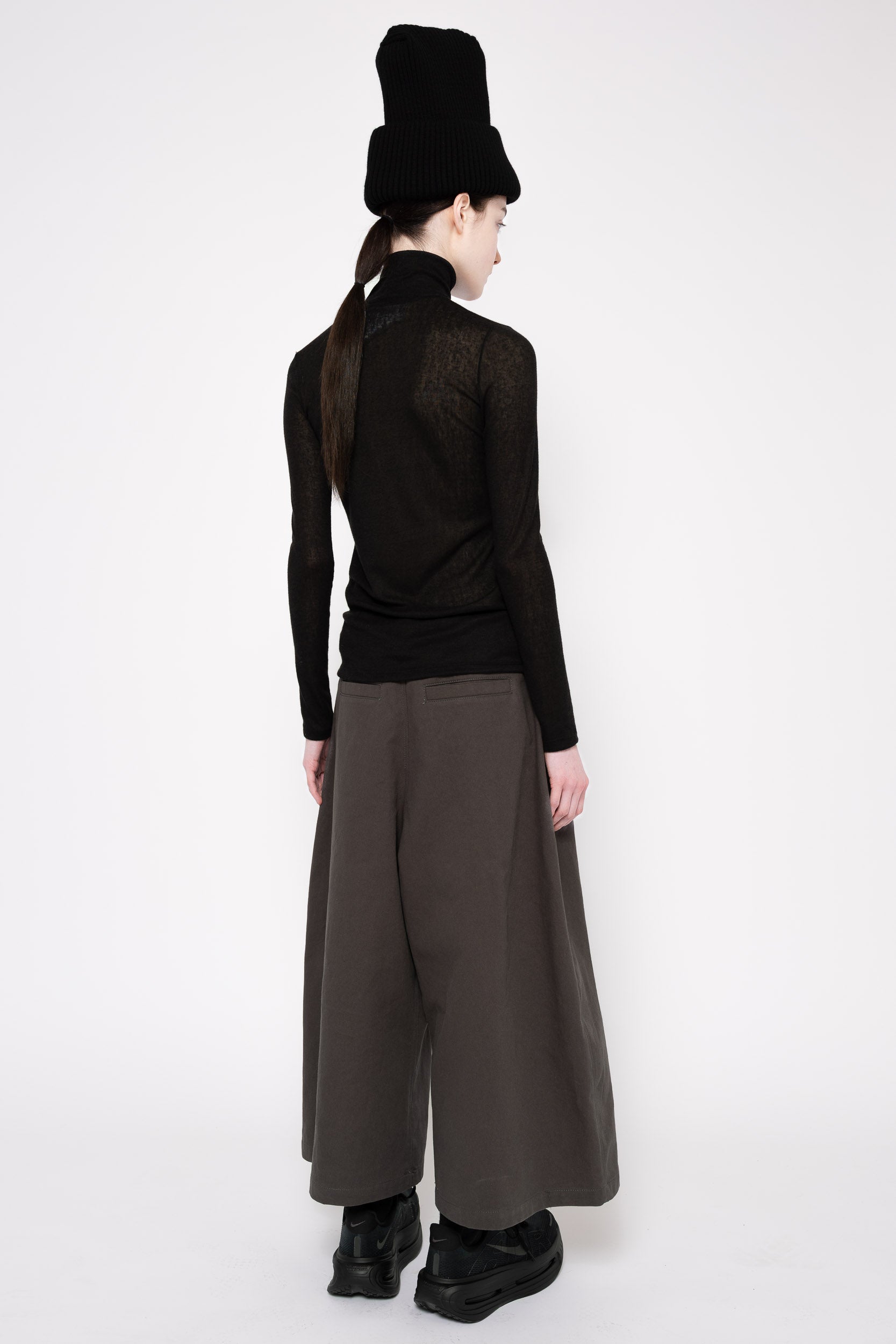 A model shown from the back wearing a black long-sleeve knit top, wide-leg dark brown trousers, a black fuzzy hat, and black boots against a plain white studio background.