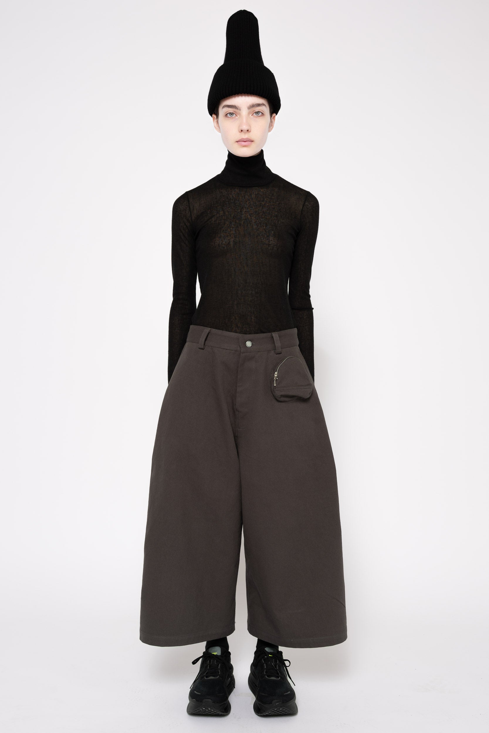 A model stands against a light gray studio background wearing a black knit turtleneck, wide-leg dark brown cropped trousers with a side pocket, a black pointed beanie, and black lace-up shoes.
