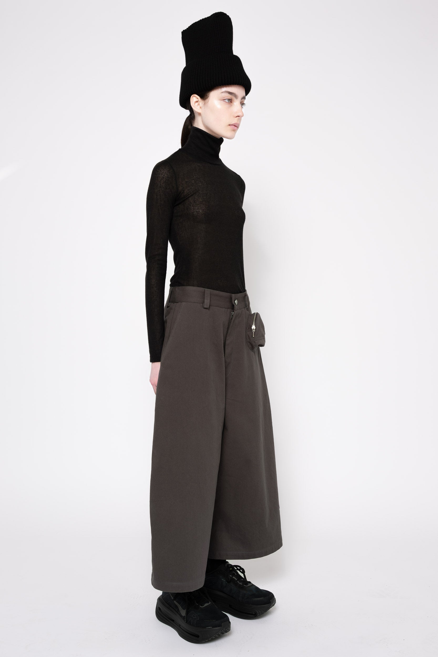 A model is shown in profile wearing a black sheer long-sleeve turtleneck top, wide-leg dark brown trousers, a black ribbed hat, and black shoes against a plain white studio background.