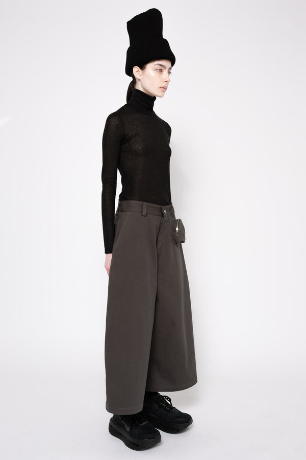 A model is shown in profile wearing a black sheer long-sleeve turtleneck top, wide-leg dark brown trousers, a black ribbed hat, and black shoes against a plain white studio background.