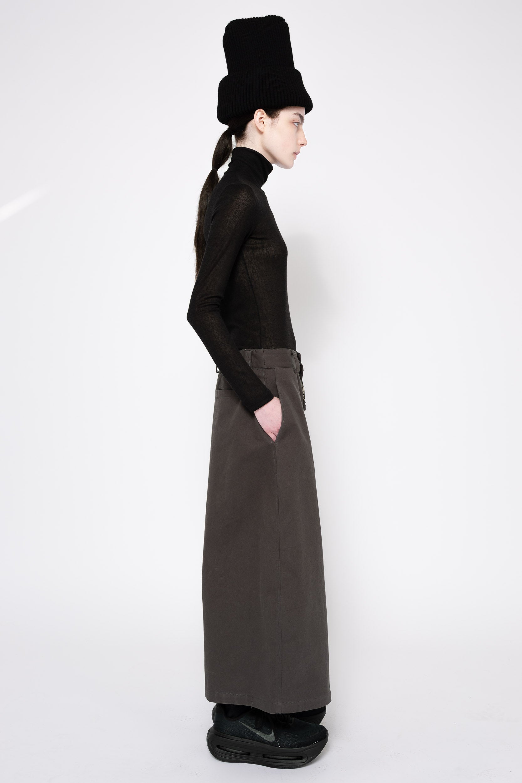 A model shown in profile wearing a black long-sleeve mock-neck top, a dark brown wide-leg skirt, a black ribbed hat, and black sneakers against a plain white studio background.