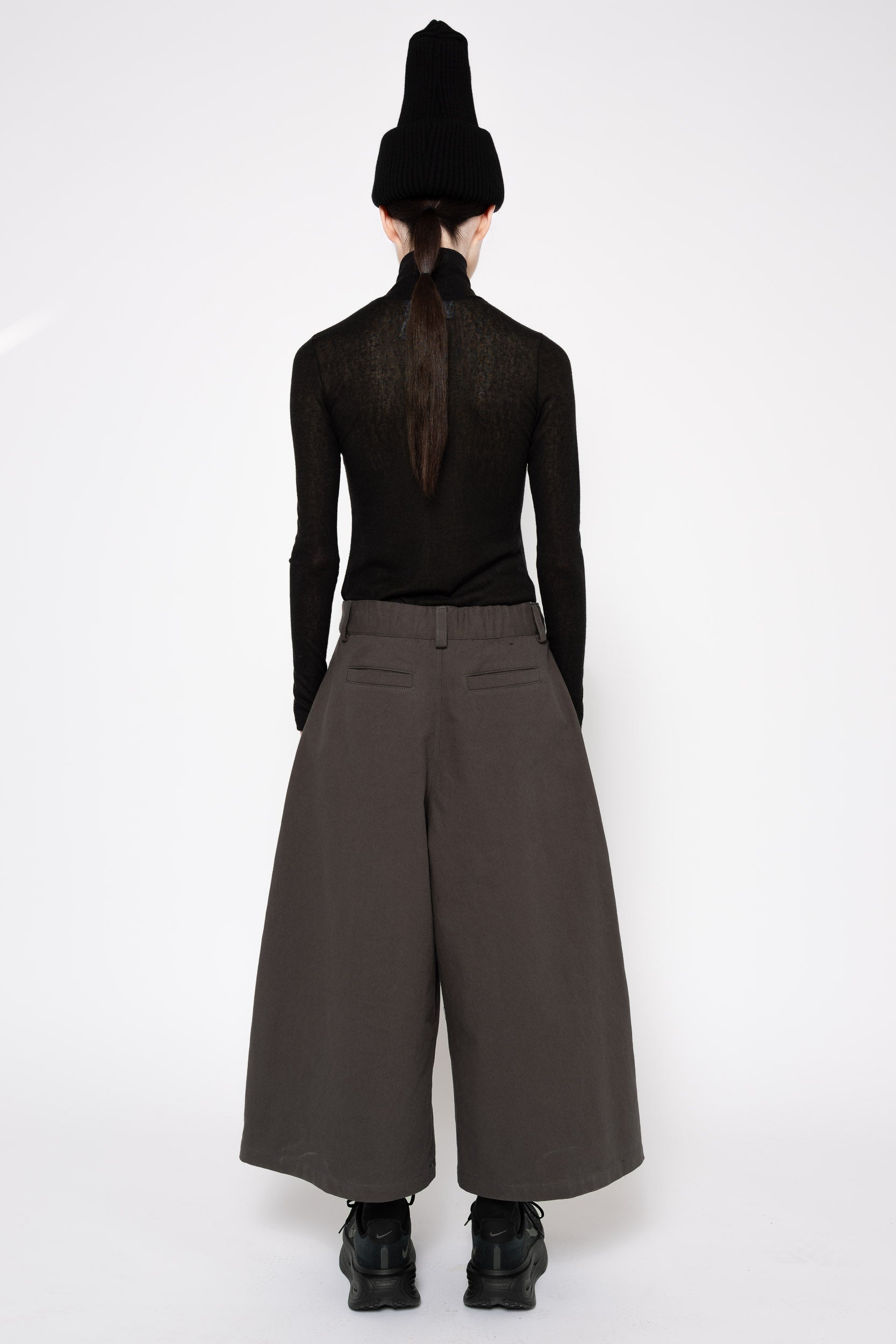 A model shown from the back wearing a black long-sleeve mesh top, wide-leg dark brown trousers, a black knit beanie, and black platform boots against a plain white studio background.