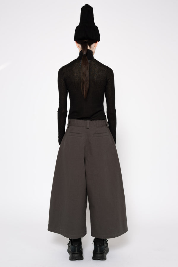 A model shown from the back wearing a black long-sleeve mesh top, wide-leg dark brown trousers, a black knit beanie, and black platform boots against a plain white studio background.