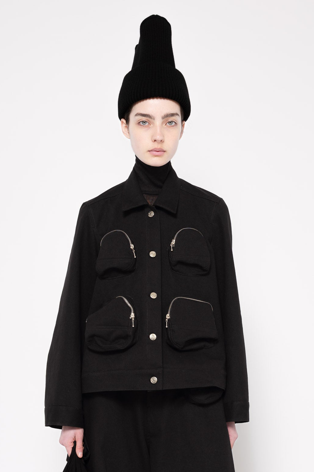A model wearing a black cropped jacket with four front pockets, a black turtleneck, and a tall black beanie against a light gray studio background.