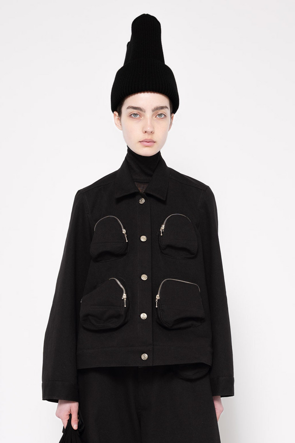 A model wearing a black cropped jacket with four front pockets, a black turtleneck, and a tall black beanie against a light gray studio background.