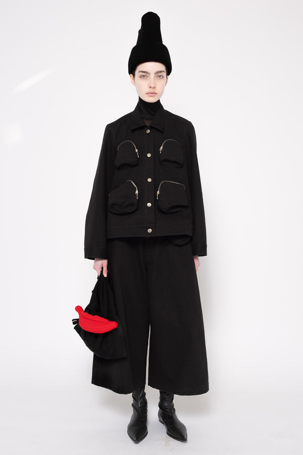 A model stands against a white studio background wearing a black high-neck jacket with multiple front pockets, black wide-leg pants, a tall black hat, and black boots, while holding a black bag with a bright red accent.