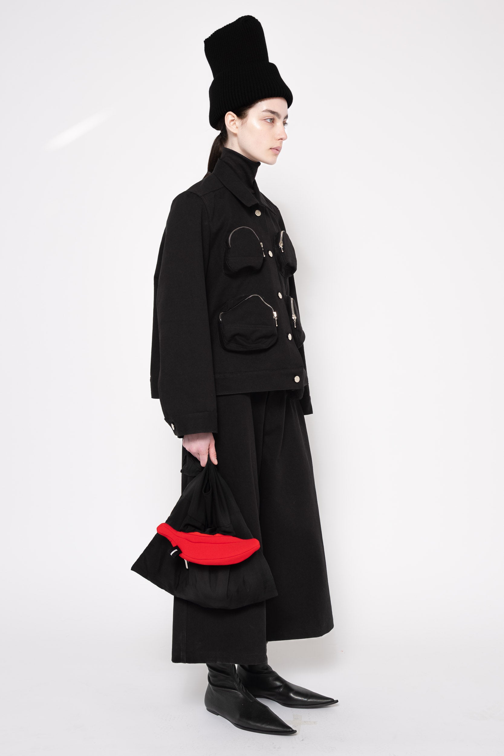 A model is shown in profile wearing a black high-collared jacket with silver hardware, a long black skirt, a black top hat, and holding a black handbag with a bold red lining, set against a plain white studio background.