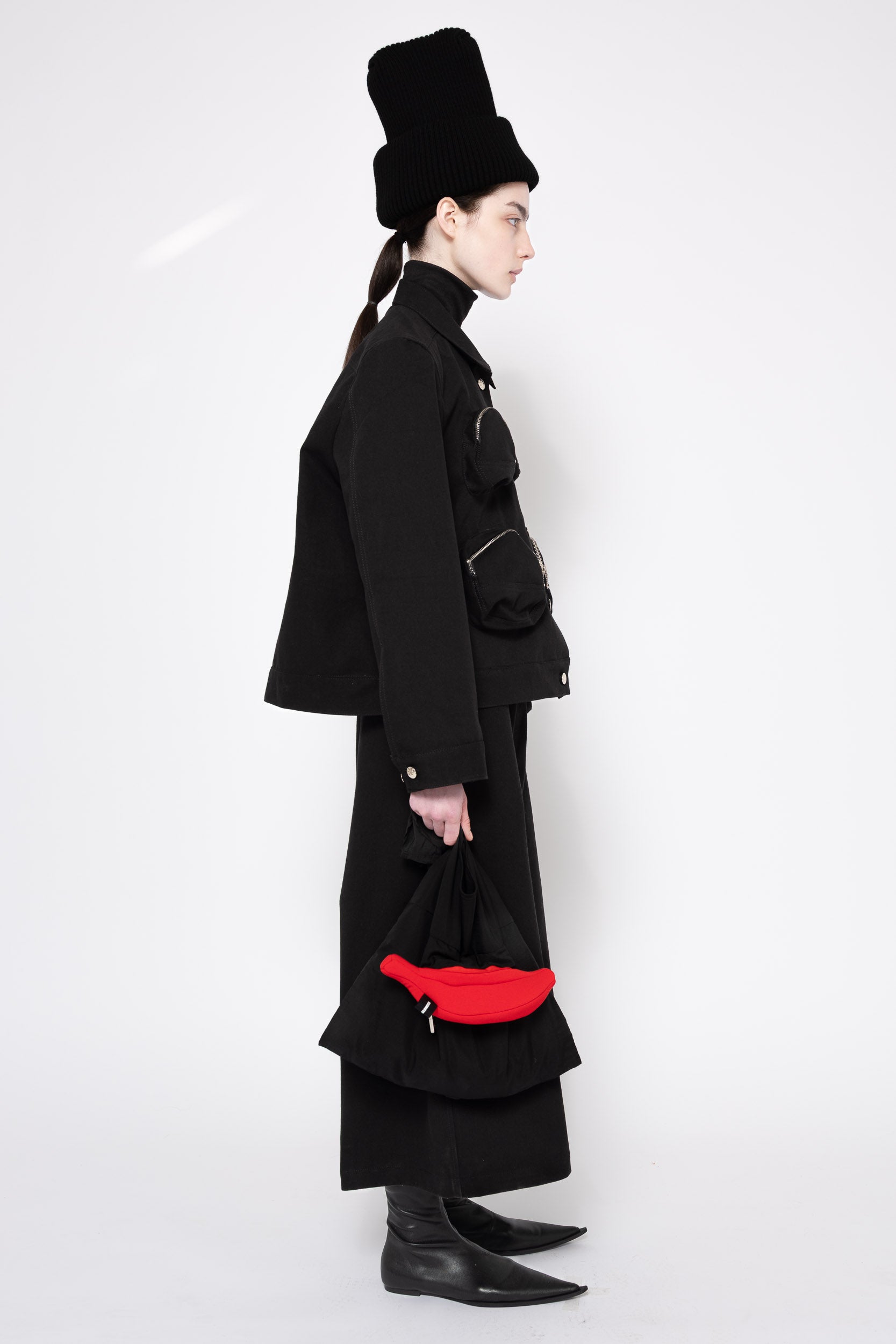 A model in profile wearing a black high-collared coat, matching wide-leg skirt, black boots, and a black top hat, holding a bright red fanny pack against a plain white studio background.