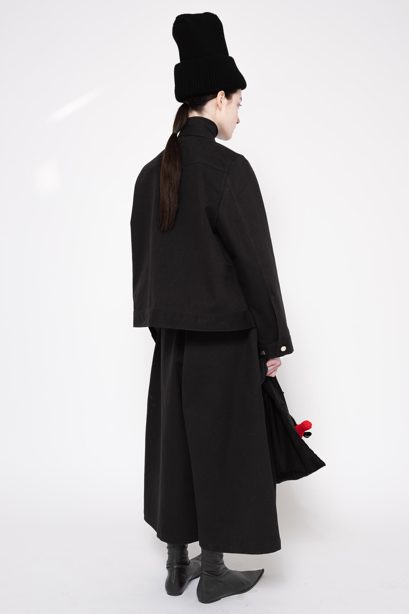 A model shown from the back wearing a black high-collared jacket, wide-leg skirt, black boots, and a black hat, holding a black handbag with a red accent against a white studio background.