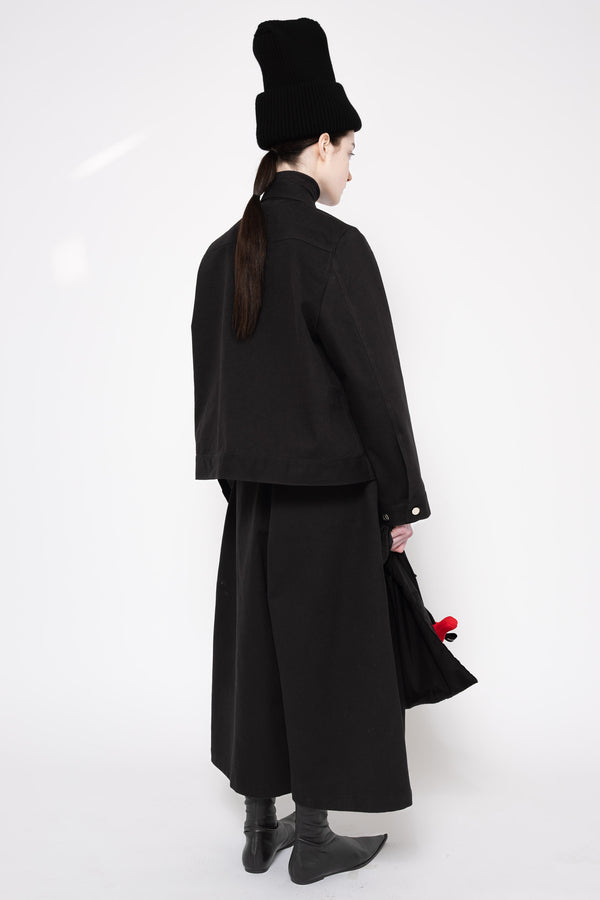 A model shown from the back wearing a black high-collared jacket, wide-leg skirt, black boots, and a black hat, holding a black handbag with a red accent against a white studio background.