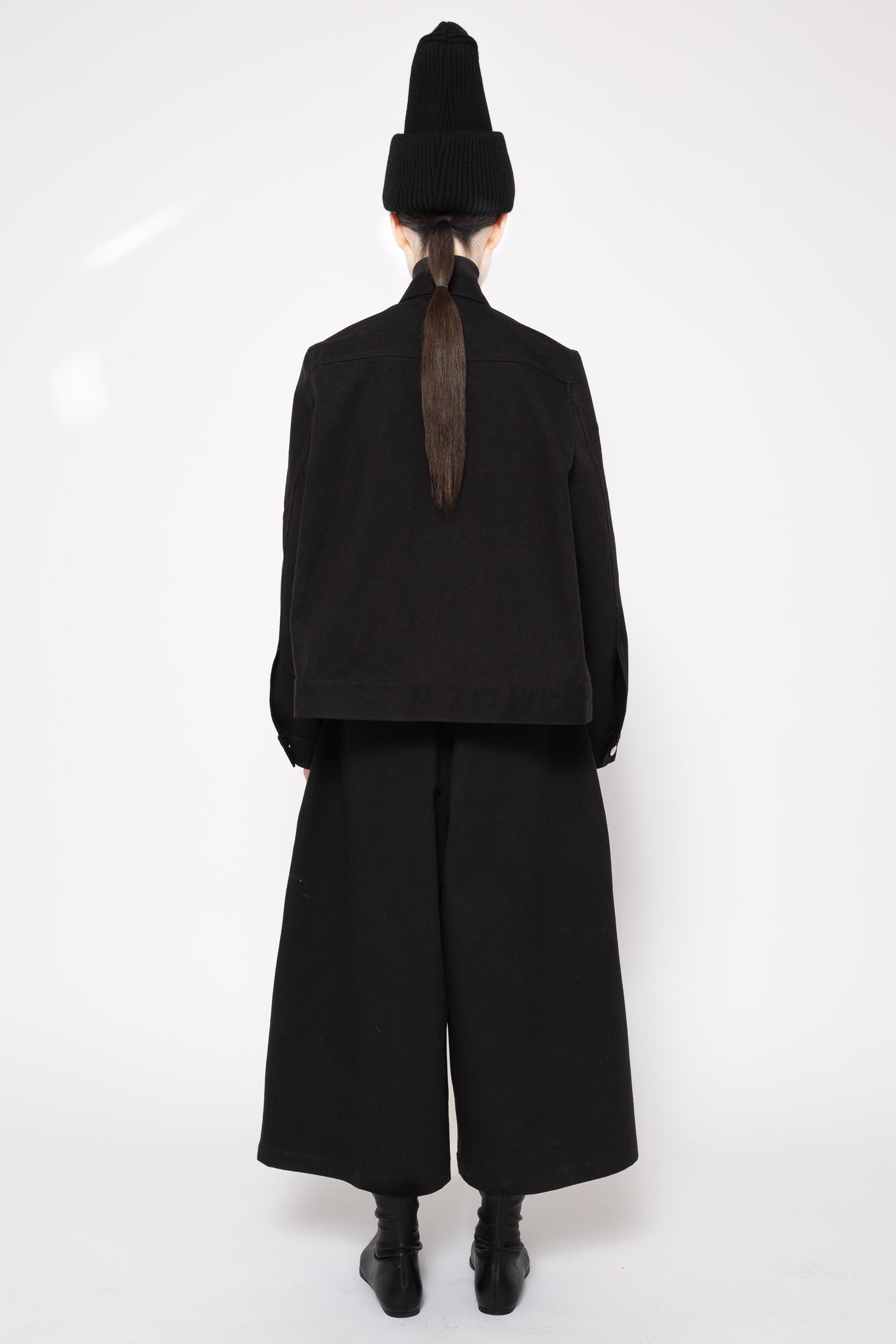 A model shown from the back wearing a black traditional-style outfit with a tall pointed hat, wide-leg pants, and black boots against a plain white studio background.