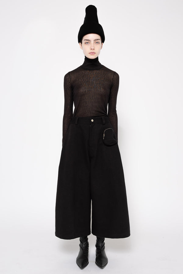 A model wearing a black sheer long-sleeve turtleneck top, wide-leg black trousers, a black beanie, and black ankle boots, standing against a plain white studio background.