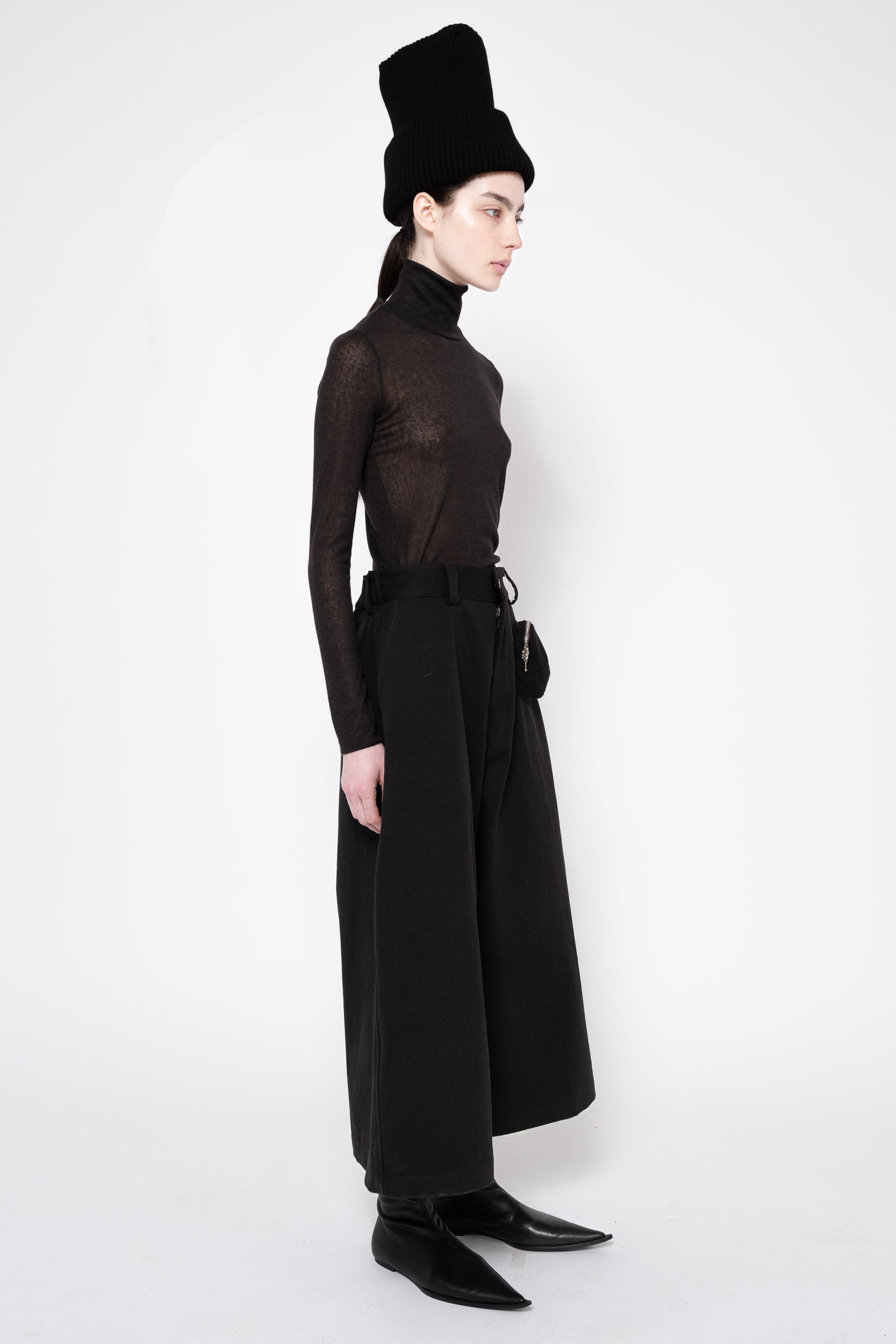 A model in profile wearing a black sheer long-sleeve turtleneck top, wide-leg black trousers, a black belt, black pointed shoes, and a tall black hat against a plain white studio background.