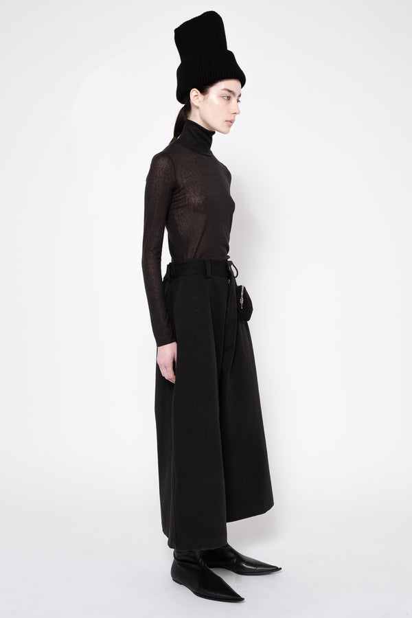 A model in profile wearing a black sheer long-sleeve turtleneck top, wide-leg black trousers, a black belt, black pointed shoes, and a tall black hat against a plain white studio background.