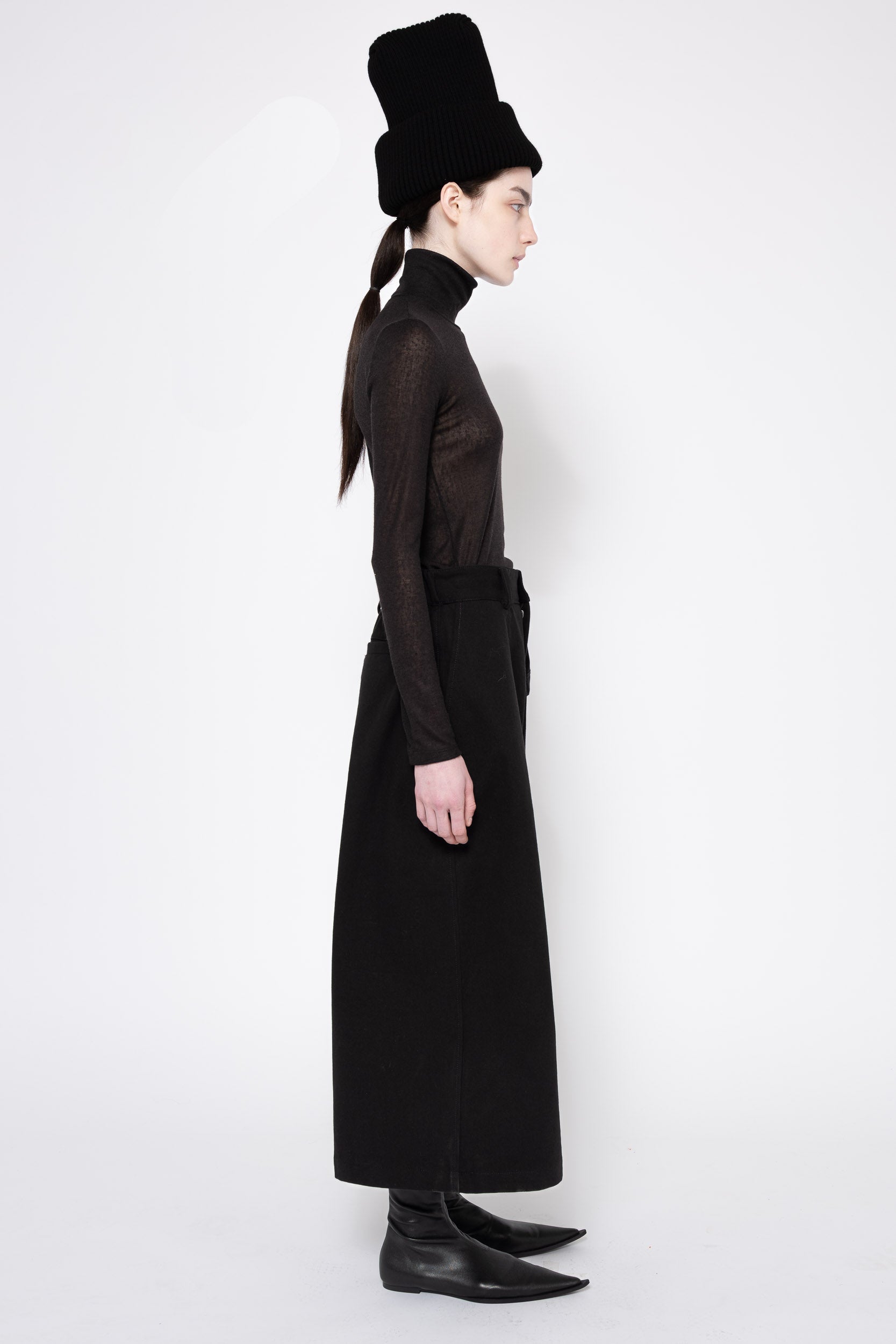 A model shown in profile wearing a long black sheer long-sleeve top with a matching black skirt and a tall black hat, set against a plain white studio background.