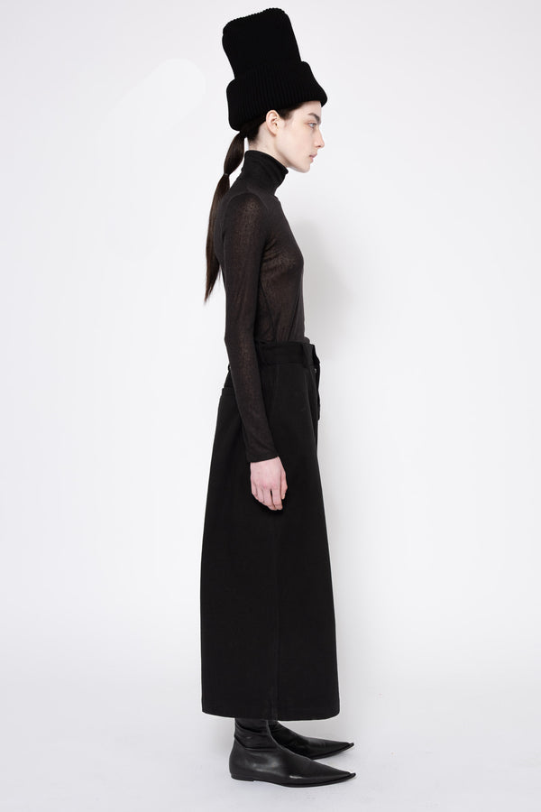 A model shown in profile wearing a long black sheer long-sleeve top with a matching black skirt and a tall black hat, set against a plain white studio background.