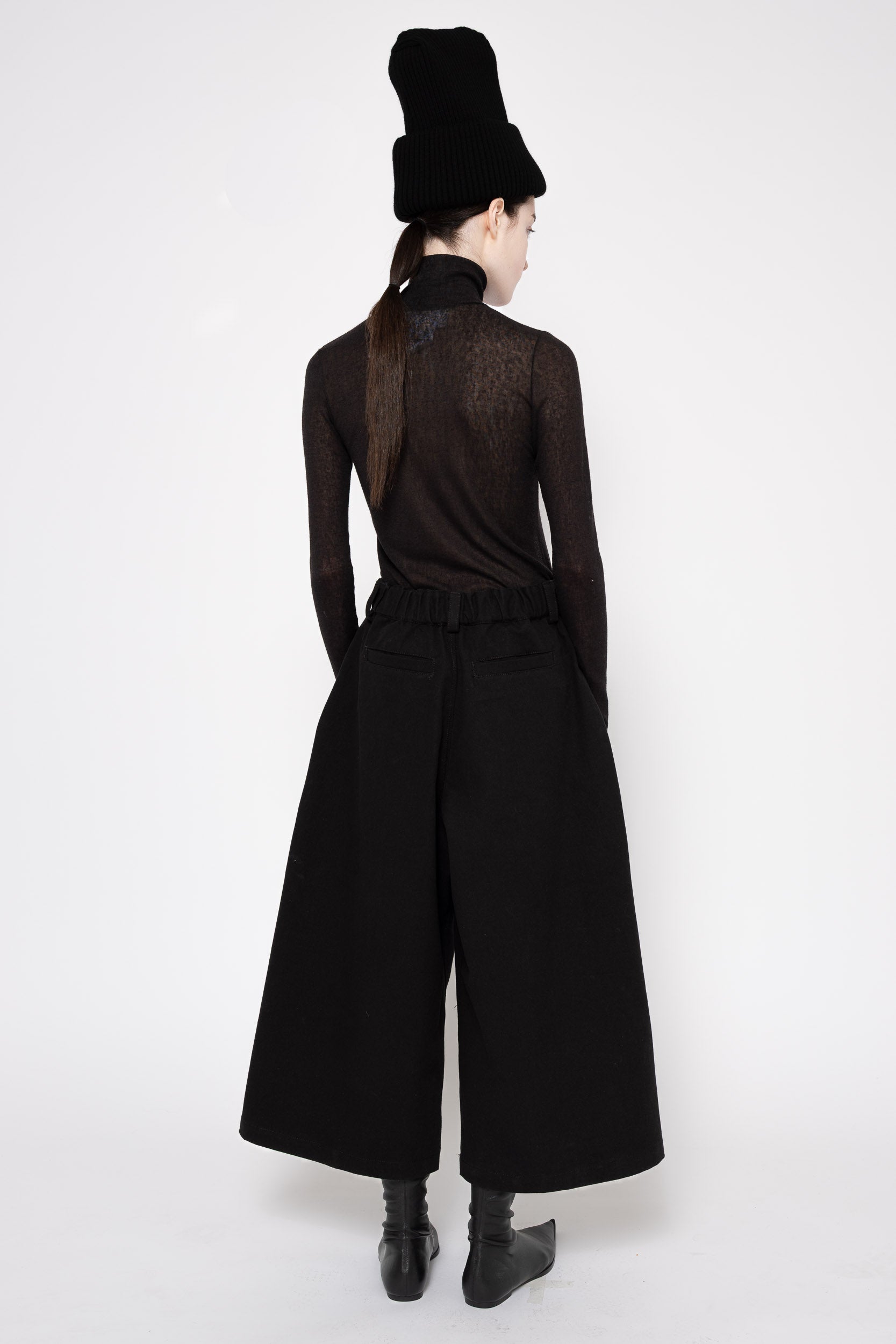 Back view of a model wearing all black turtleneck with wide leg culottes and a high knit black hat. Knee high black boots.