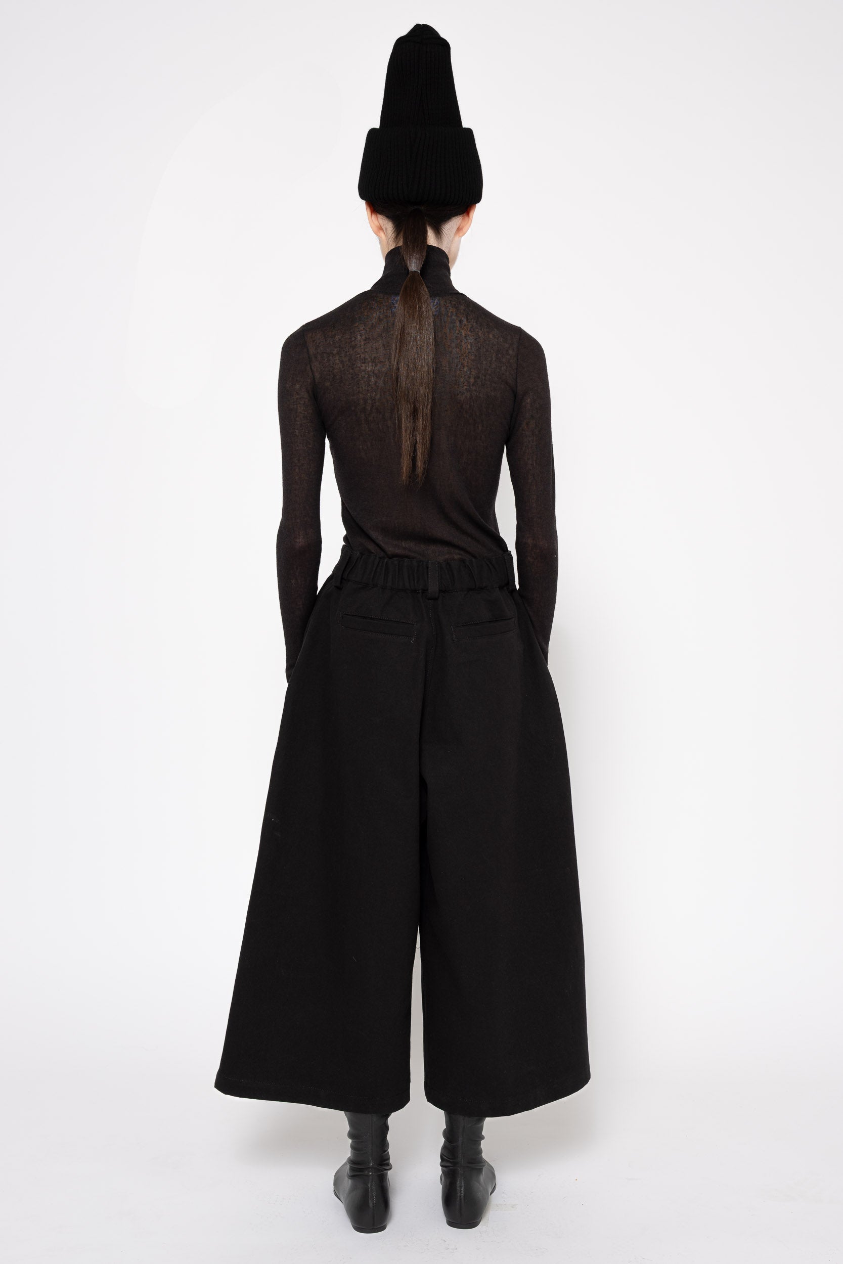 Back view of a model wearing all black turtleneck with wide leg culottes and a high knit black hat. Knee high black boots.