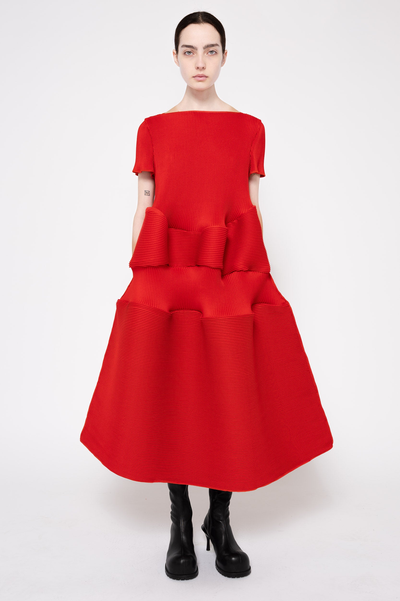 A model wearing a bright red off-the-shoulder midi dress with structured, tiered ruffle details on the skirt and sleeves, paired with black ankle boots, against a plain white studio background.