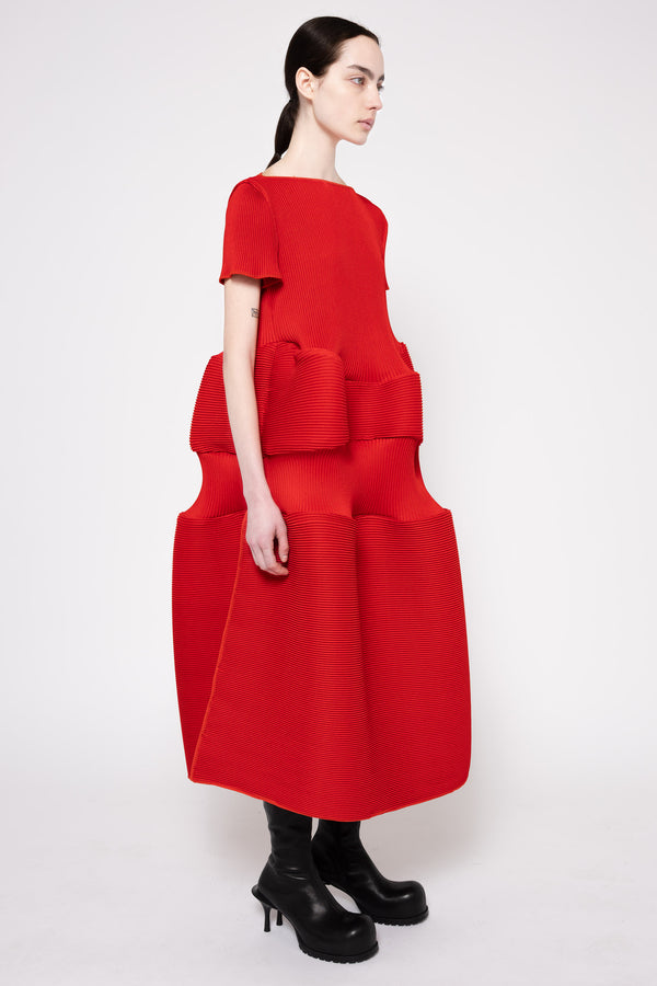 A model wearing a bright red midi dress with a unique layered, boxy texture and black leather boots, photographed against a plain light gray studio background.
