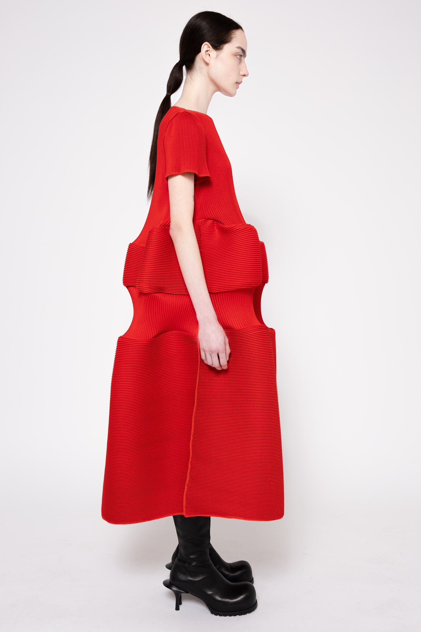 A model shown from the side profile wearing a bright red, sculptural midi dress with layered ruffles and short sleeves, paired with black platform boots, against a plain light gray studio background.