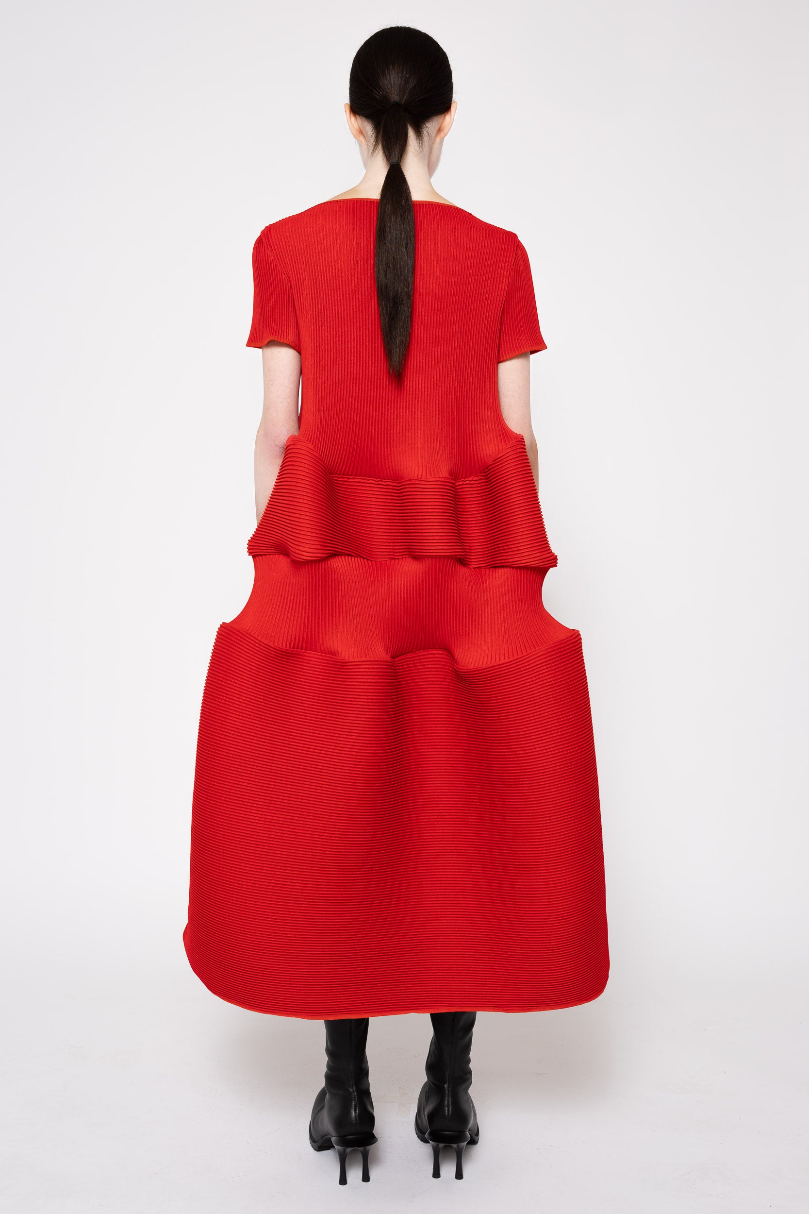 A model shown from the back wearing a bright red short-sleeve dress with a structured, tiered ruffled waist and a flared skirt, paired with black high-heeled boots against a light gray studio background.