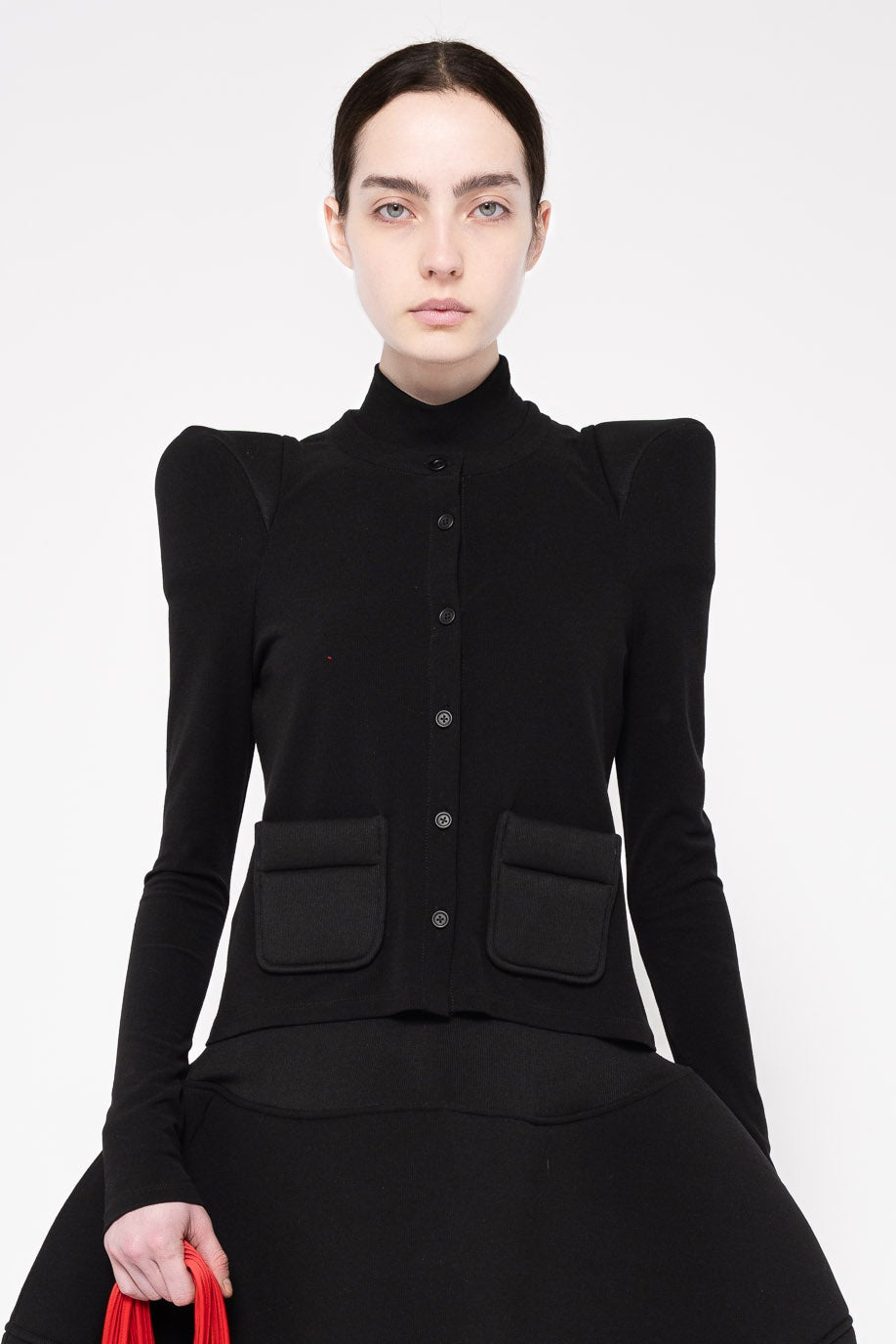 A model wearing a black long-sleeve button-front jacket with dramatic puffed shoulders and two large front patch pockets, paired with a black skirt, against a plain light gray studio background.