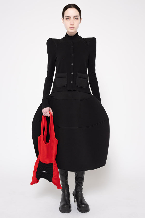 A model stands against a light gray studio background wearing a black structured midi dress with dramatic shoulder pads, a high mock neck, front buttons, and large front pockets. She is also wearing black knee-high boots and holding a bright red tote bag.