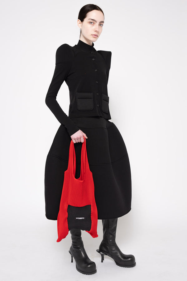 A model wearing a black structured long-sleeve jacket with puffed shoulders, a matching black skirt, and black boots, holding a bright red tote bag with a black pocket against a light gray studio background.