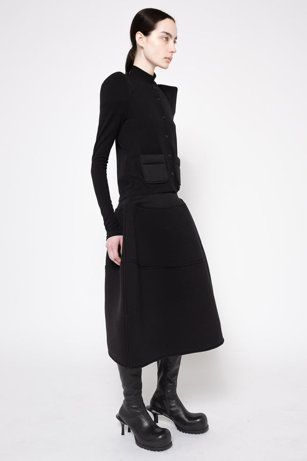 A model wearing a black long-sleeve midi dress with a structured, slightly asymmetrical collar and front patch pocket, styled with black leather boots against a plain white studio background.
