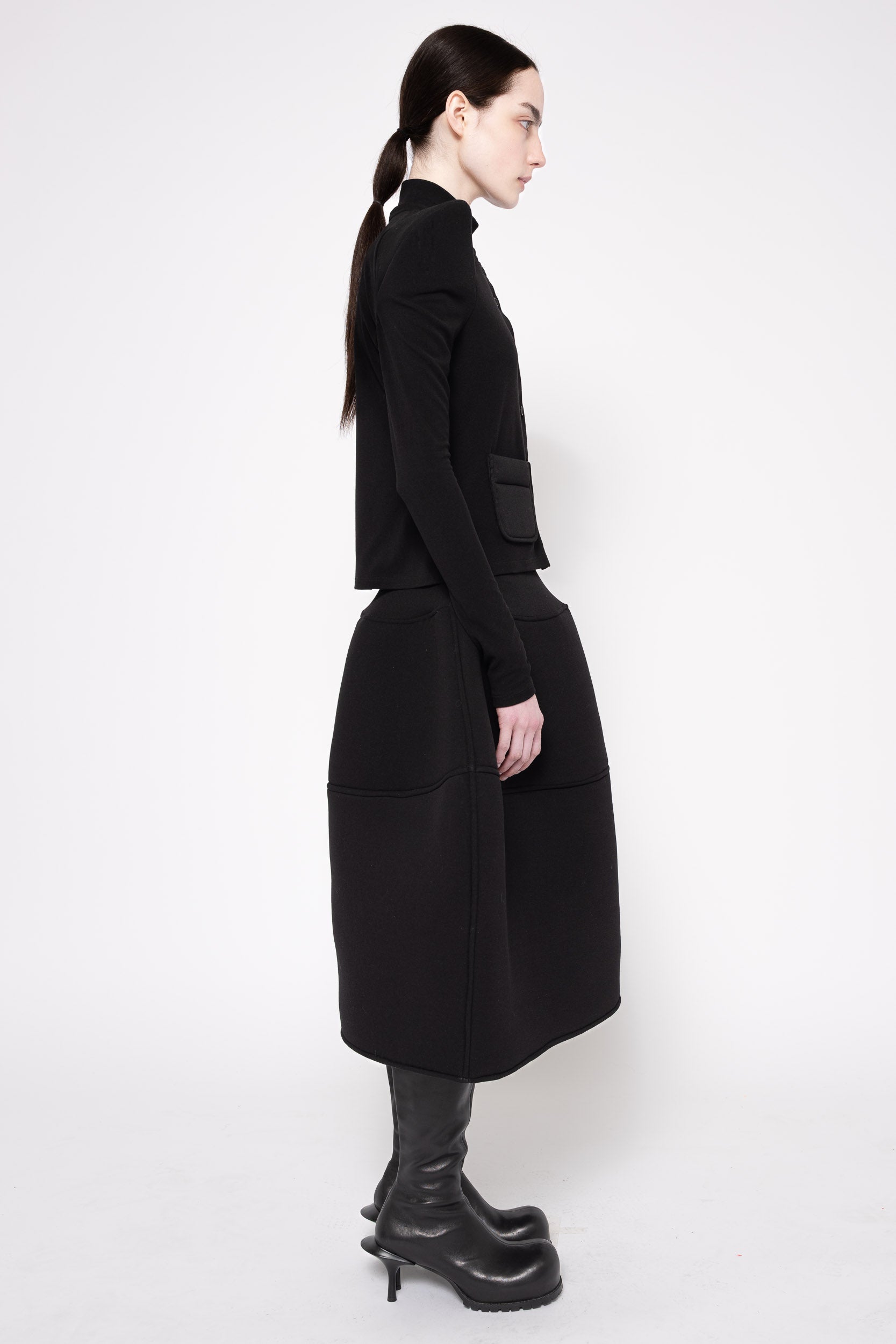 A model shown in profile wearing a black long-sleeve mock-neck top and a matching black midi skirt with large patch pockets, paired with black platform boots, against a plain white studio background.