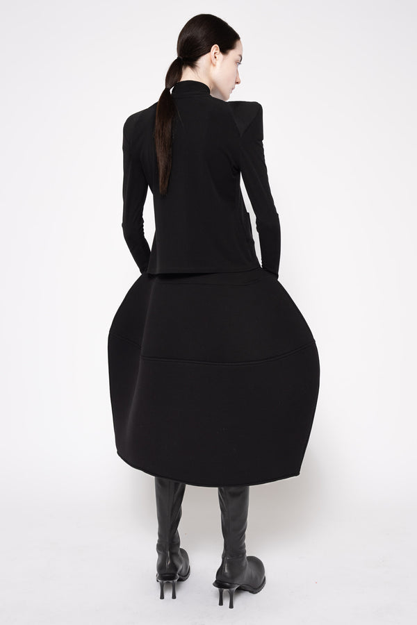 A model shown from the back wearing a black long-sleeve dress with dramatic shoulder detailing and a voluminous skirt, paired with black pointed-toe heels, against a plain white studio background.