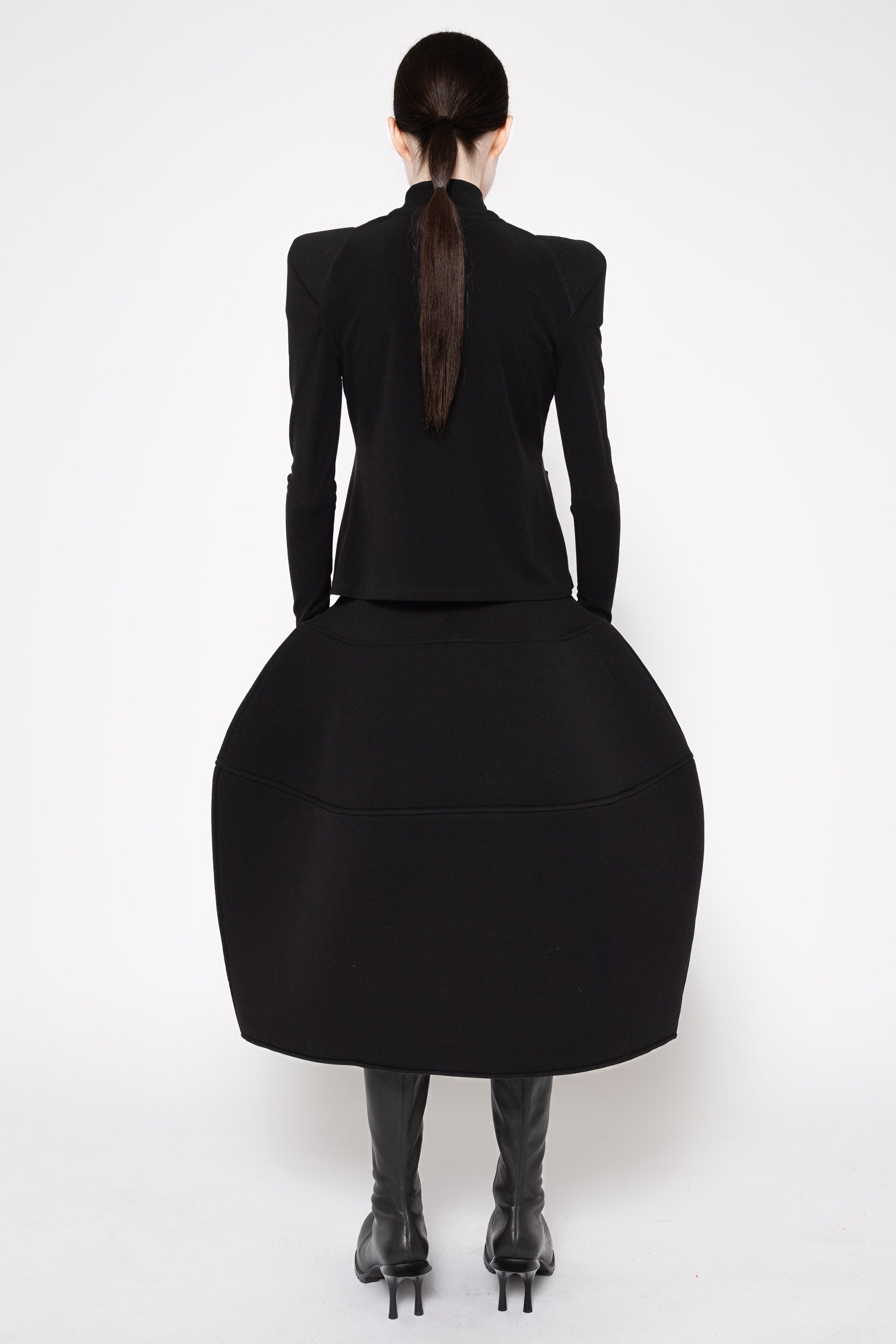 A model shown from the back wearing a black structured long-sleeve top with a dramatic, oversized flared skirt and black knee-high boots, set against a plain white studio background.