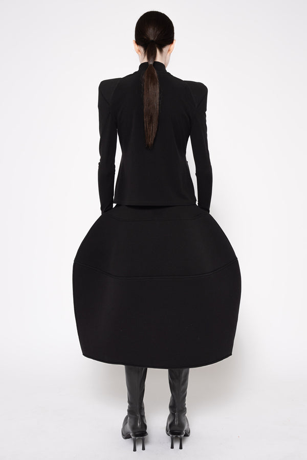 A model shown from the back wearing a black structured long-sleeve top with a dramatic, oversized flared skirt and black knee-high boots, set against a plain white studio background.