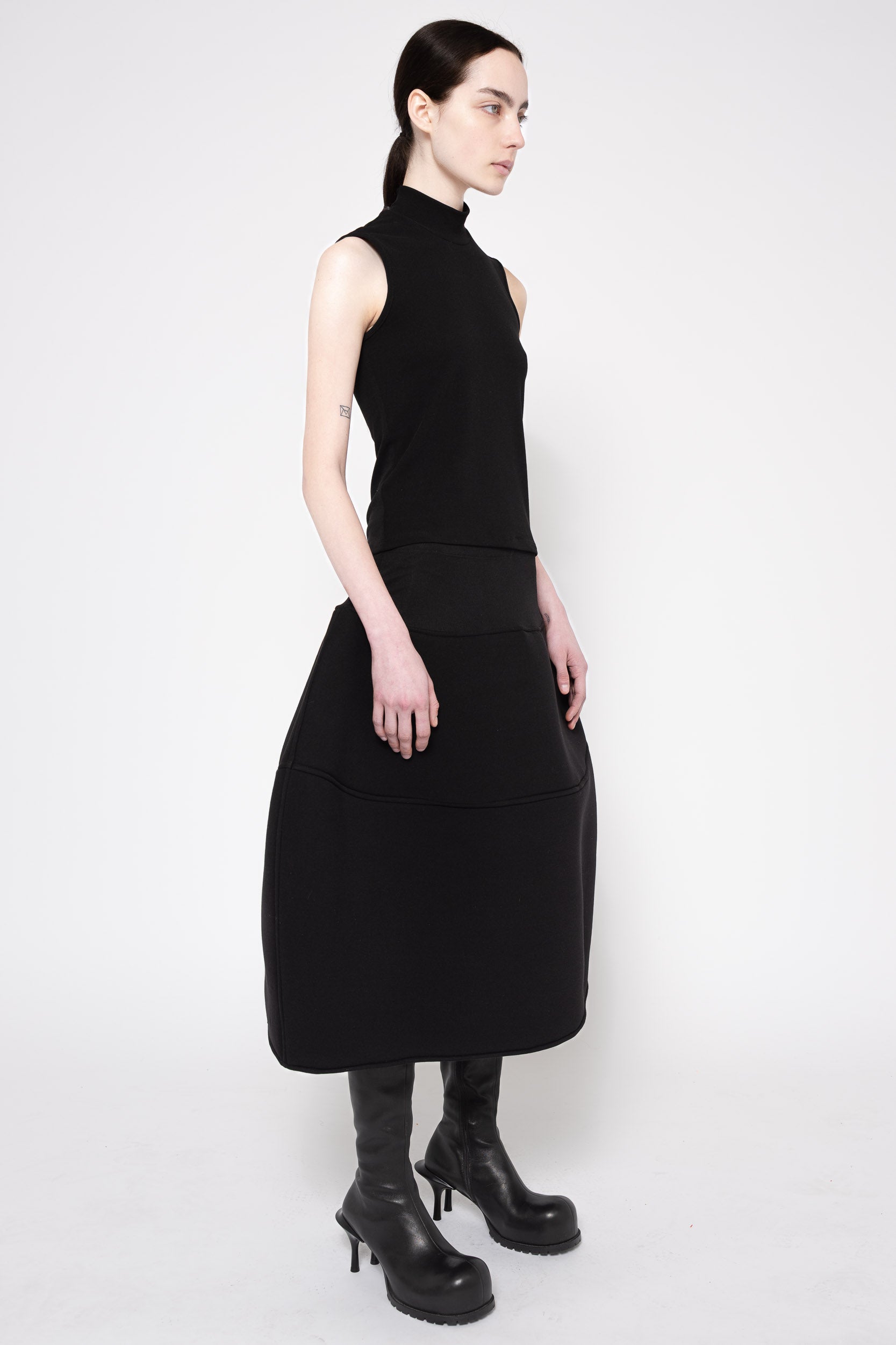 A model wearing a sleeveless black mock-neck midi dress with a wide waistband and black leather boots, photographed against a plain light gray studio background.