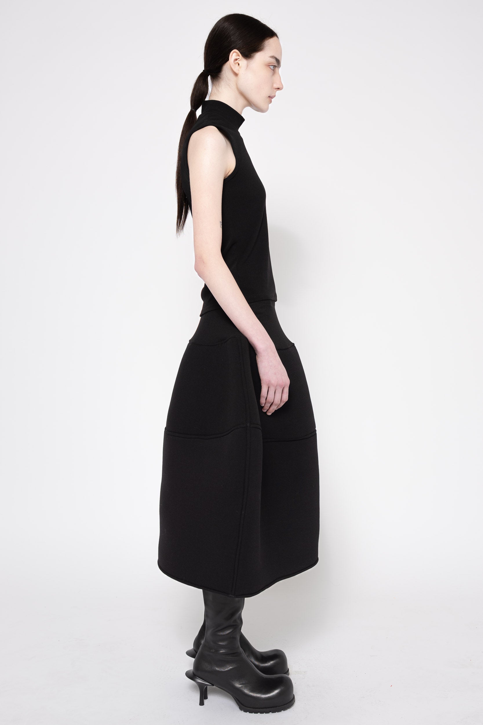 A model in profile wearing a sleeveless black high-neck midi dress with a structured, voluminous skirt and black leather boots, photographed against a plain light gray studio background.