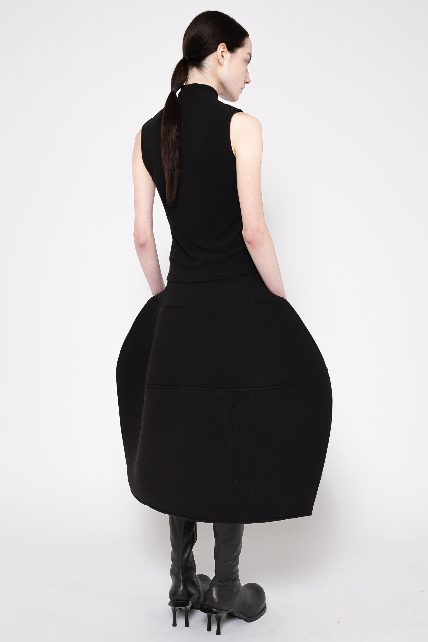 A model shown from the back wearing a sleeveless black midi dress with a high mock neck and a voluminous, balloon-like skirt, paired with black boots, against a plain white studio background.