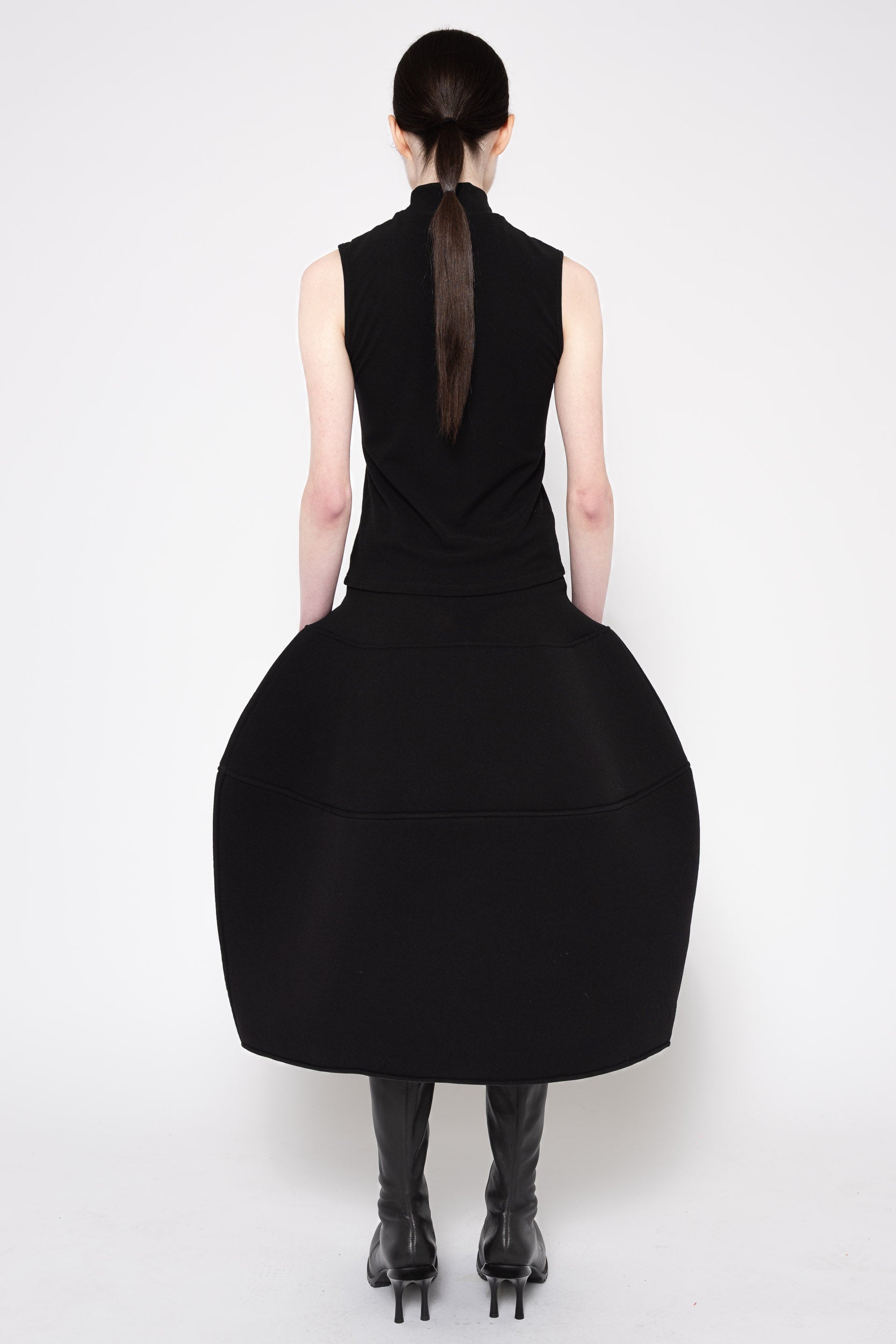 A model shown from the back wearing a black sleeveless dress with a high neck and a very large, voluminous skirt, paired with black knee-high boots against a plain white studio background.